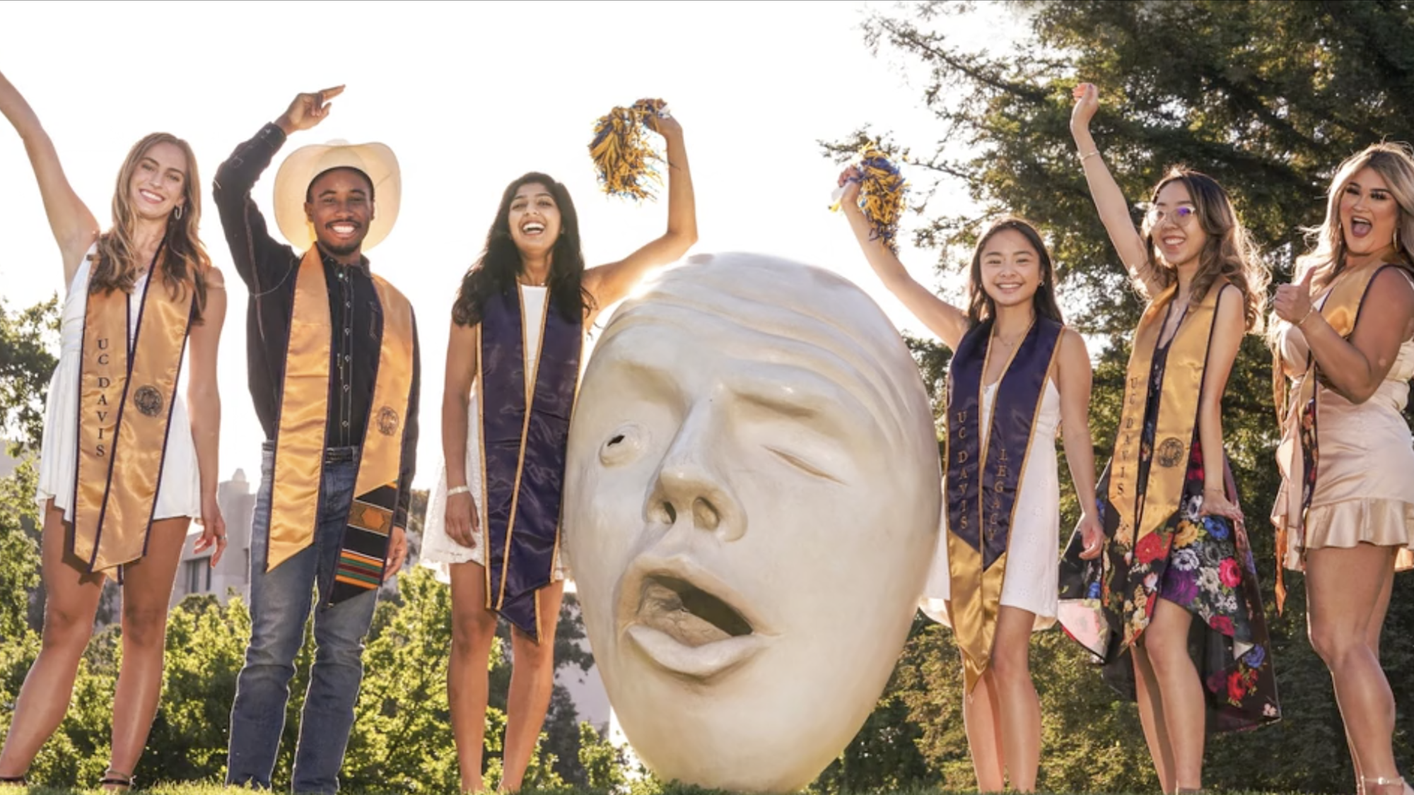 six students in graduation hats and sashes pose near an Egghead statue at TV