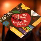 A grad cap that reads, "And the student became the teacher," in white cursive within a bright red apple, surrounded by teaching equipment like rulers, crayons and a pencil that reads, "Ms. Murillo." The photo was taken at TV' School of Education Commencement graduation ceremony in 2023.