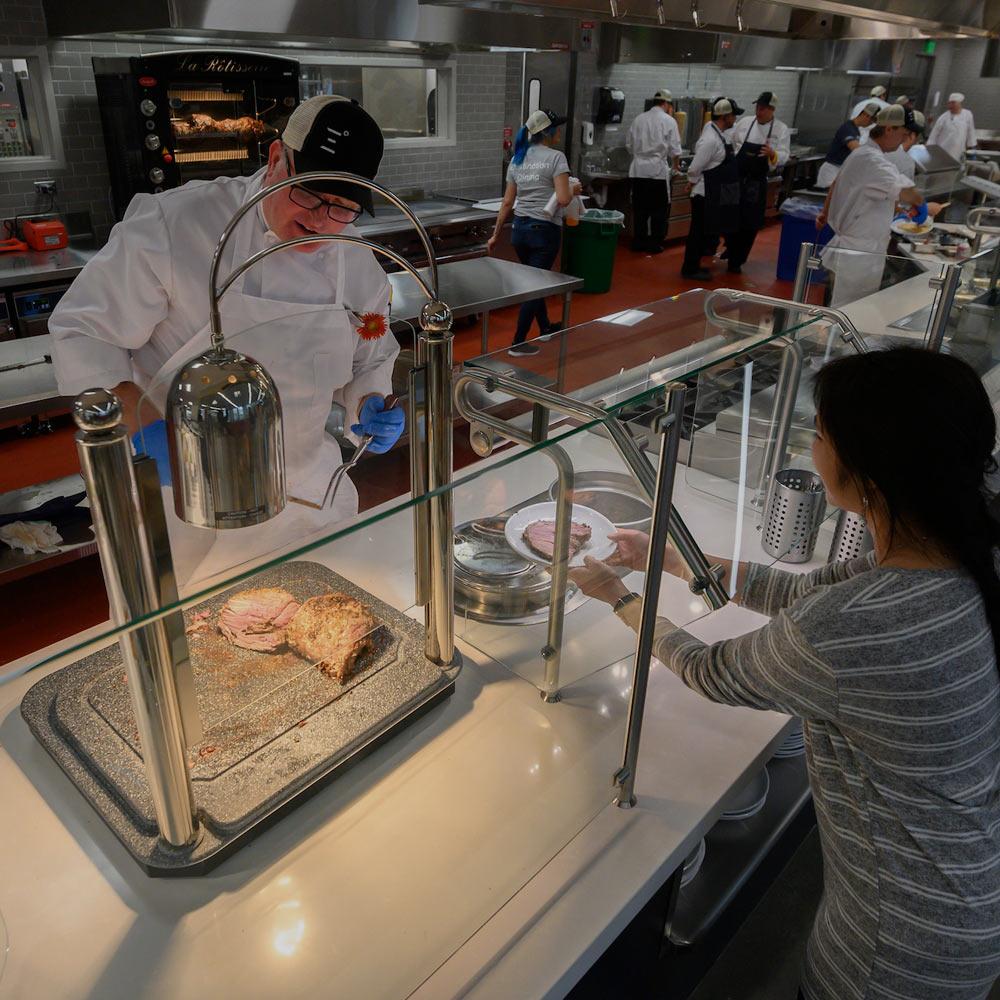 A dining commons employee serves a cut of meat to a TV student