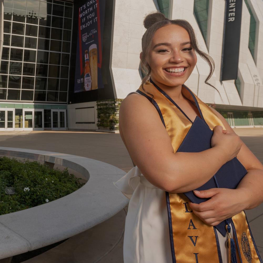 A TV student standing outside of the Golden 1 center holdint their grad regalia